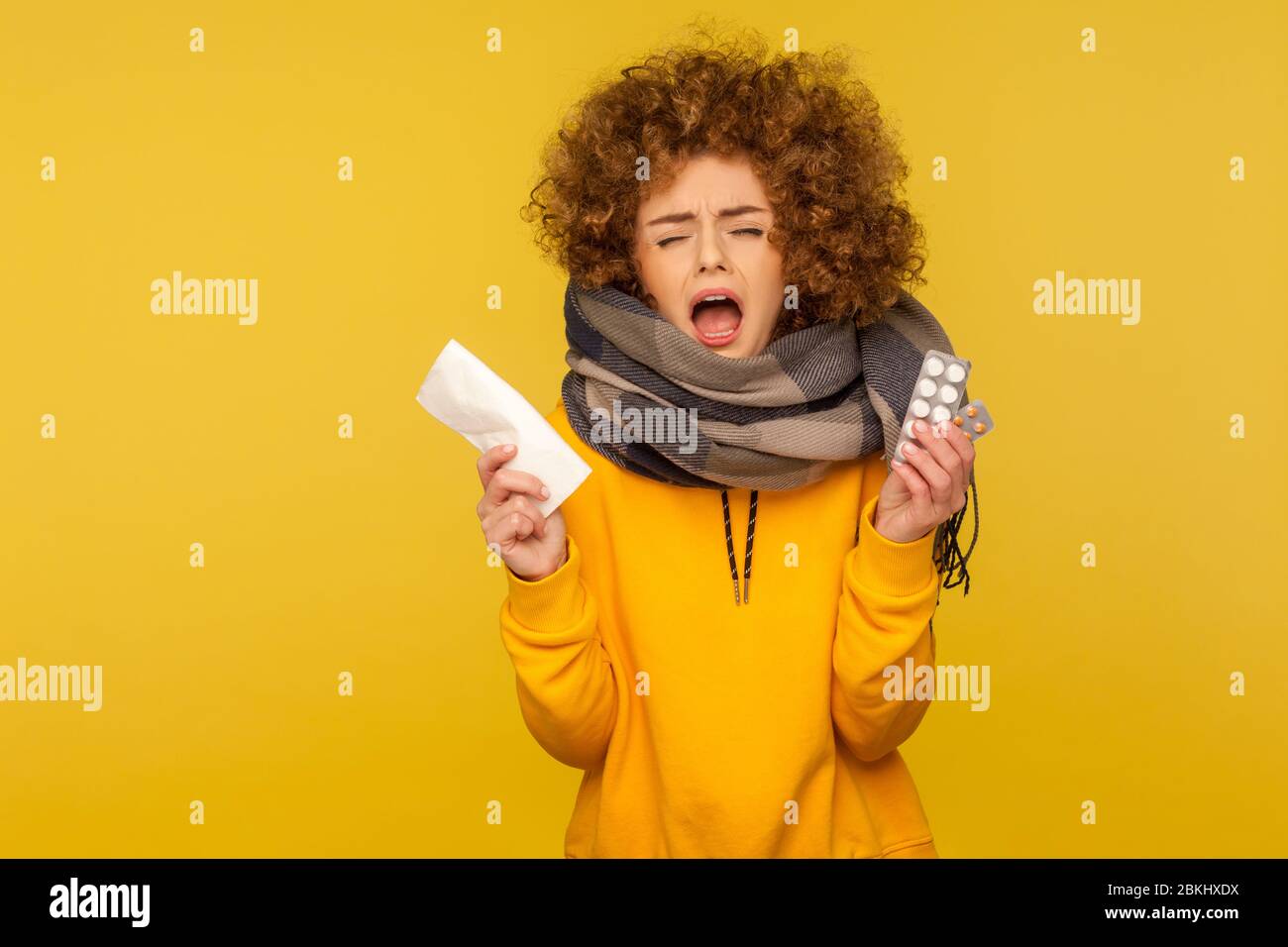 Traitement de la grippe saisonnière. Femme malade de la grippe avec des cheveux bouclés molletonnés, enveloppés dans une écharpe chaude, éternuant et tenant une serviette avec des pilules de médicaments, feein Banque D'Images