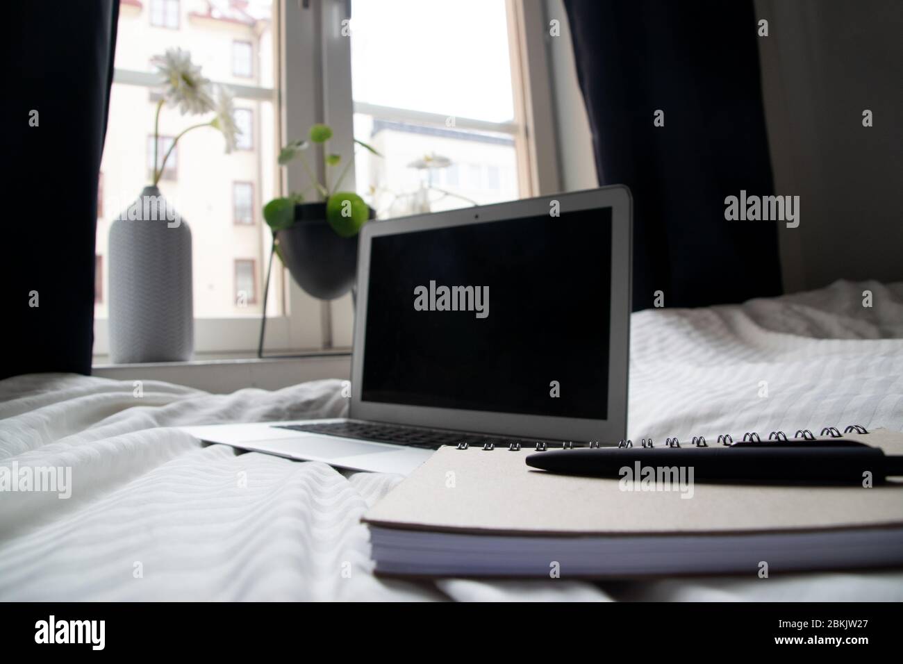 Utilisation de la chambre comme bureau à la maison avec le lit comme espace de travail. Gros plan sur le bloc-notes, le stylo et l'ordinateur portable sur le lit avec fenêtre et plantes en arrière-plan. Banque D'Images