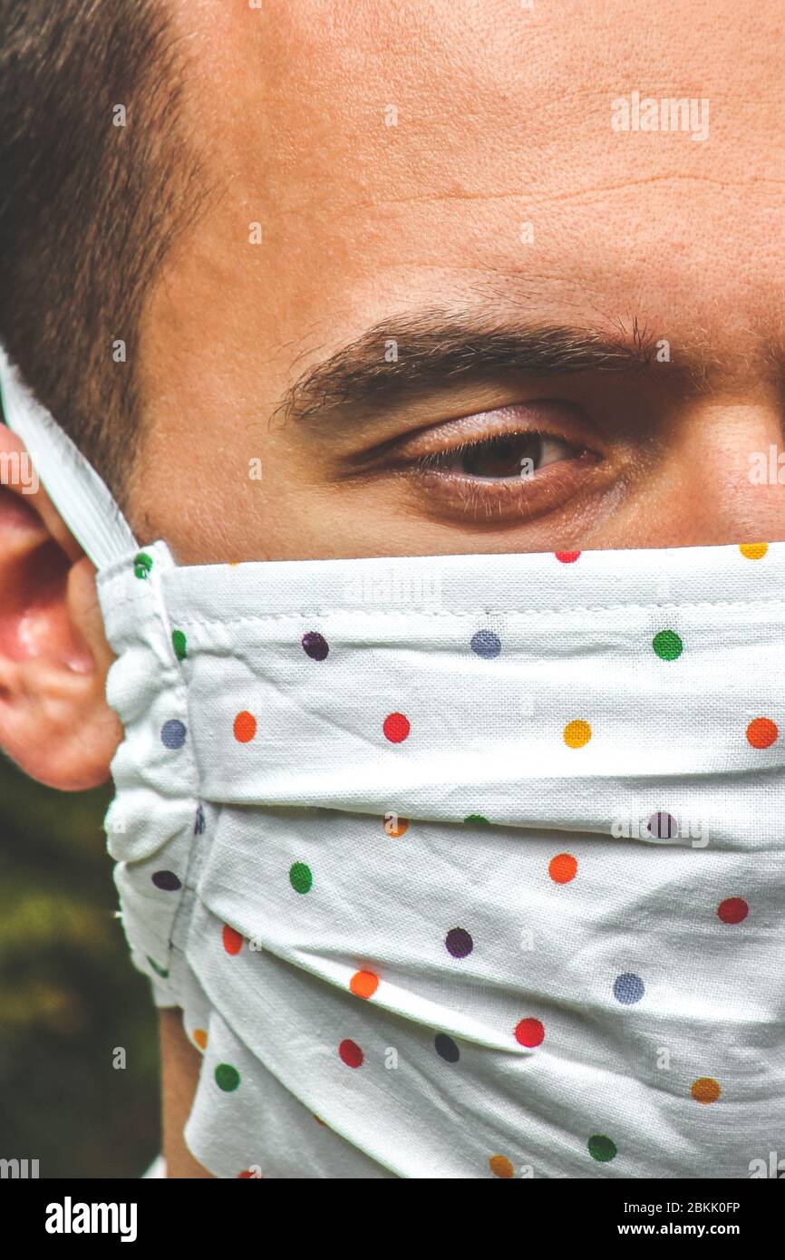 Jeune homme caucasien avec un masque de visage fait main en tissu pendant la pandémie de coronavirus. Coronavirus, éclosion de COVID-19. Concept de protection antivirus. Arrière-plan flou. Détail visage et yeux. Photo verticale. Banque D'Images