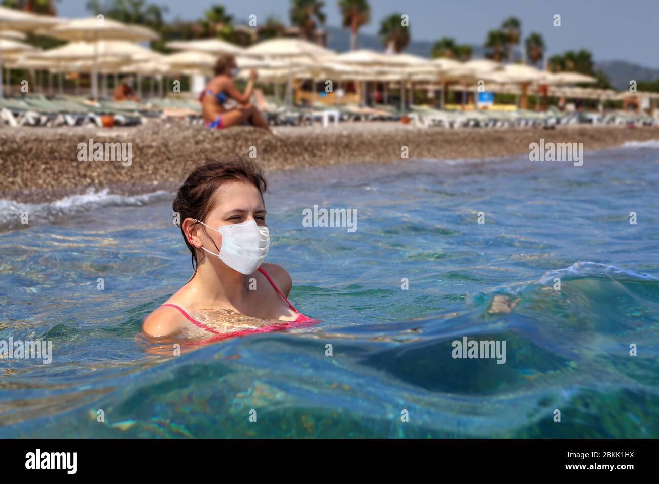 La jeune femme caucasienne nage en mer avec un masque médical protecteur,  sur fond de littoral avec parasols de plage le jour d'été ensoleillé.  Vacances dans Photo Stock - Alamy