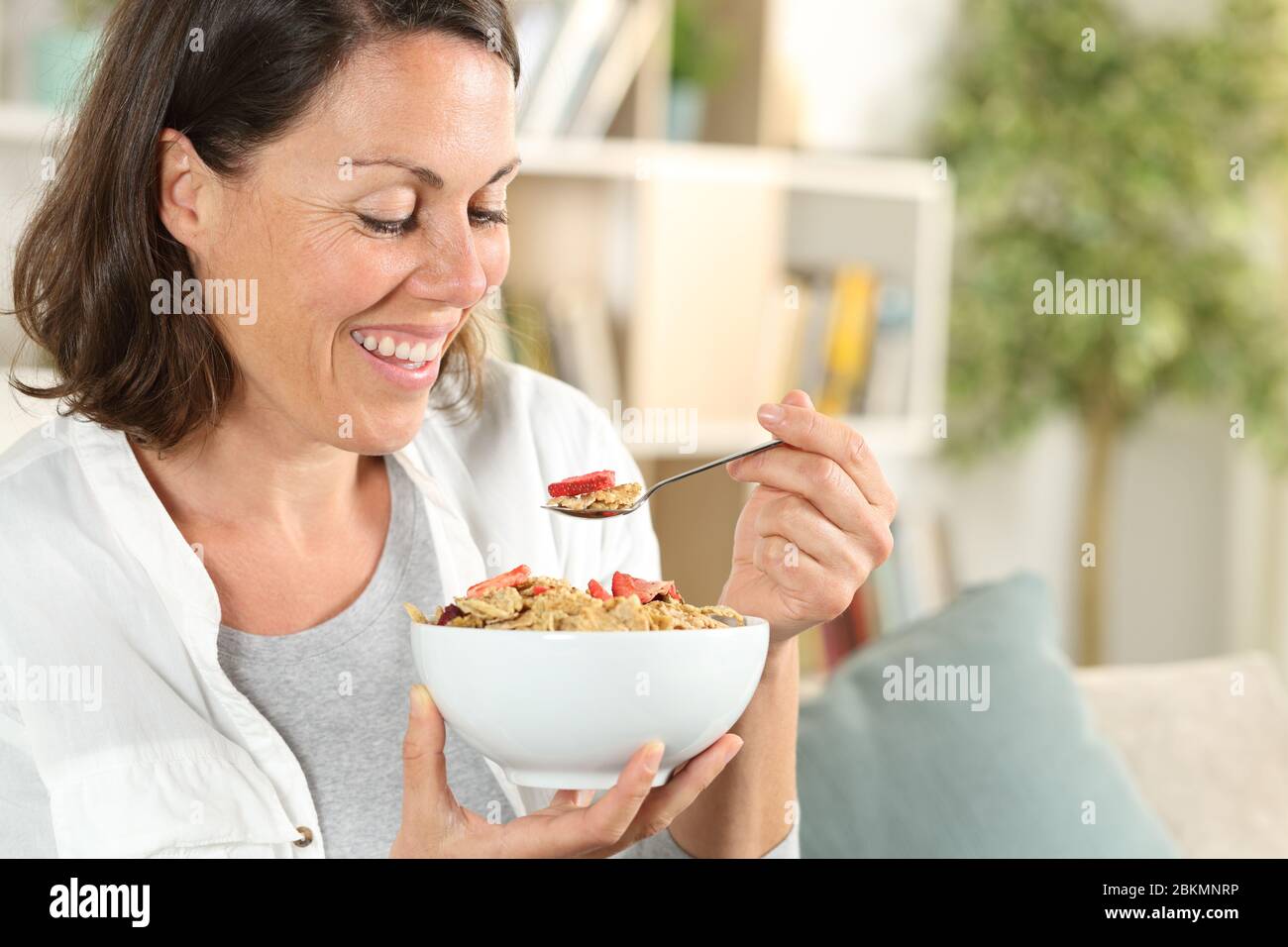 Bonne femme adulte mangeant un bol de céréales prenant le petit déjeuner assis dans le canapé à la maison Banque D'Images