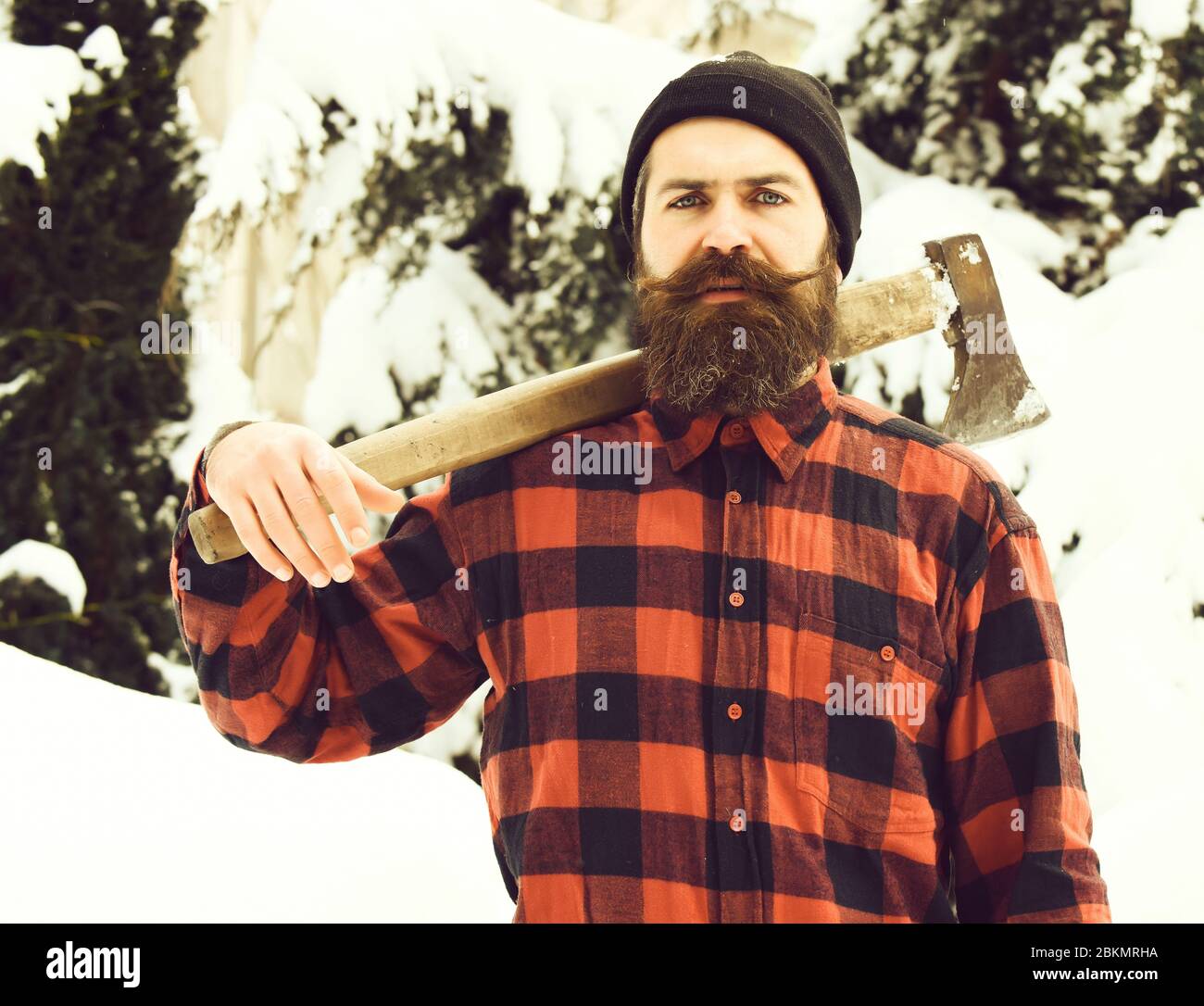 Bel homme ou bûcheron, hipster barbu, avec barbe et moustache dans des  supports de chemise à damier rouge avec hache dans la forêt enneigée le  jour de l'hiver à l'extérieur sur fond