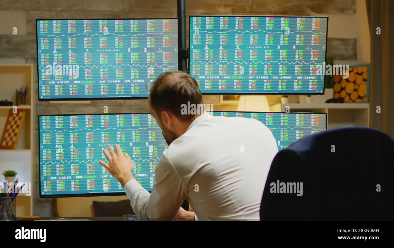 Trader barbu dans son bureau à domicile portant un costume vérifiant le marché boursier sur quatre écrans Banque D'Images