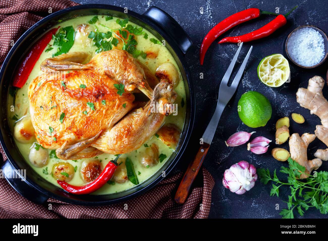 Poulet rôti au lait de coco thaïlandais et pommes de terre pour bébés au piment, feuilles de citron vert kaffir dans un braiser noir sur une table de cuisine avec fourchette à viande et ingrédients Banque D'Images