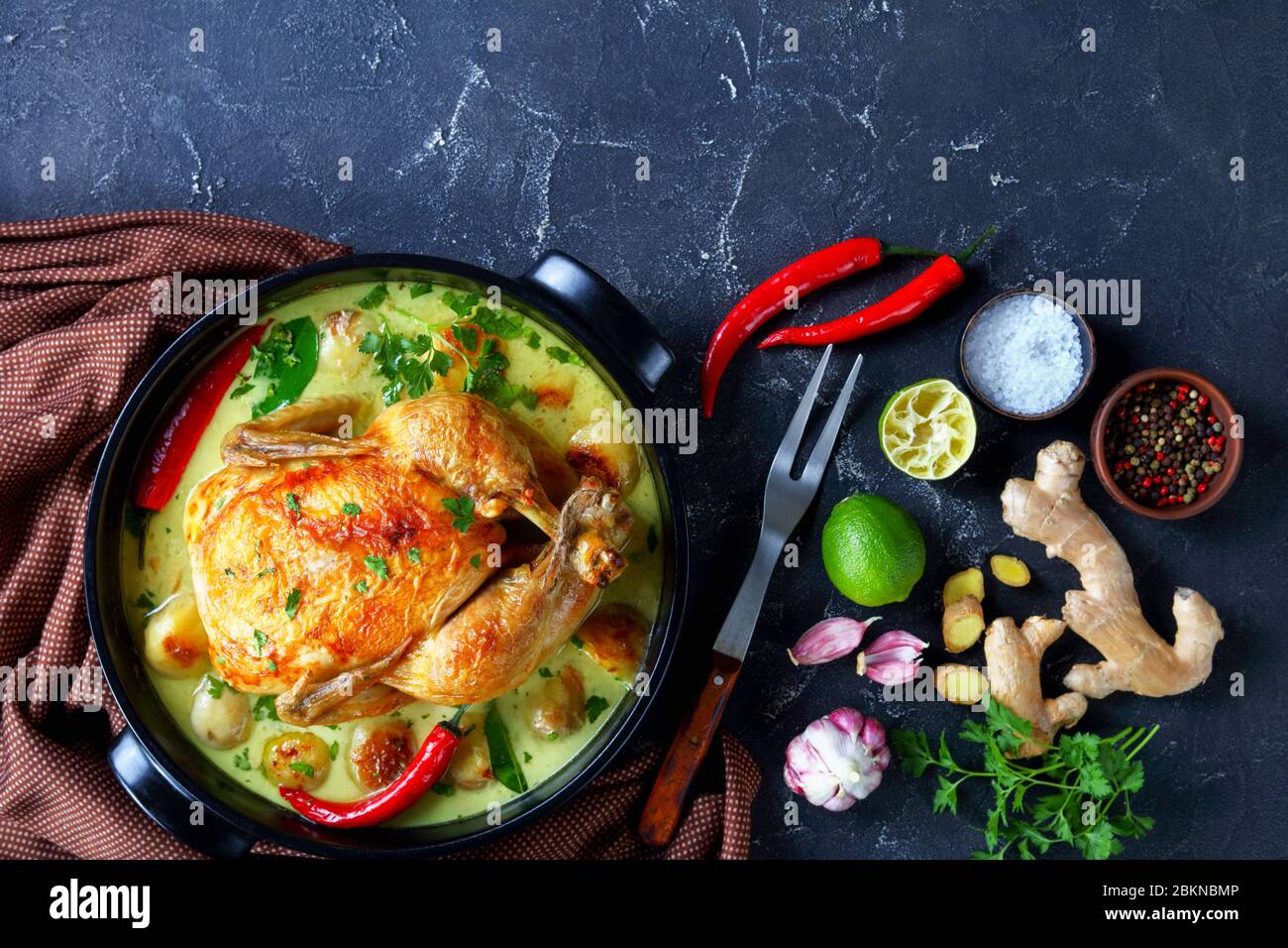 Poulet rôti au lait de coco thaïlandais et pommes de terre pour bébés au piment, feuilles de citron vert kaffir dans un braiser noir sur une table de cuisine avec fourchette à viande et ingrédients Banque D'Images