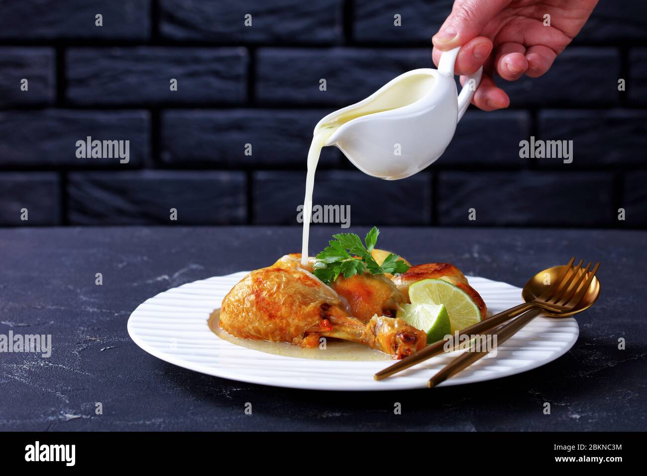 La main de femme verse de la sauce à la noix de coco goutte à goutte sur une jambe de poulet rôtie, servie avec des pommes de terre de bébé sur une assiette blanche, vue horizontale depuis le dessus Banque D'Images