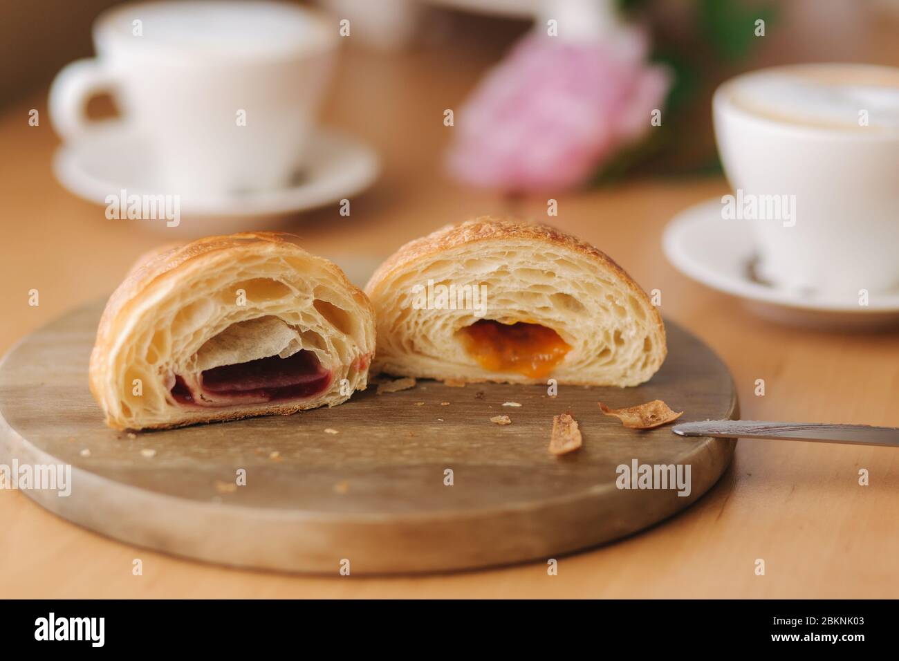 Deux moitiés de croissant fraîchement poêlé sur une planche de bois avec une confiture différente. Fond de cappuccino Banque D'Images