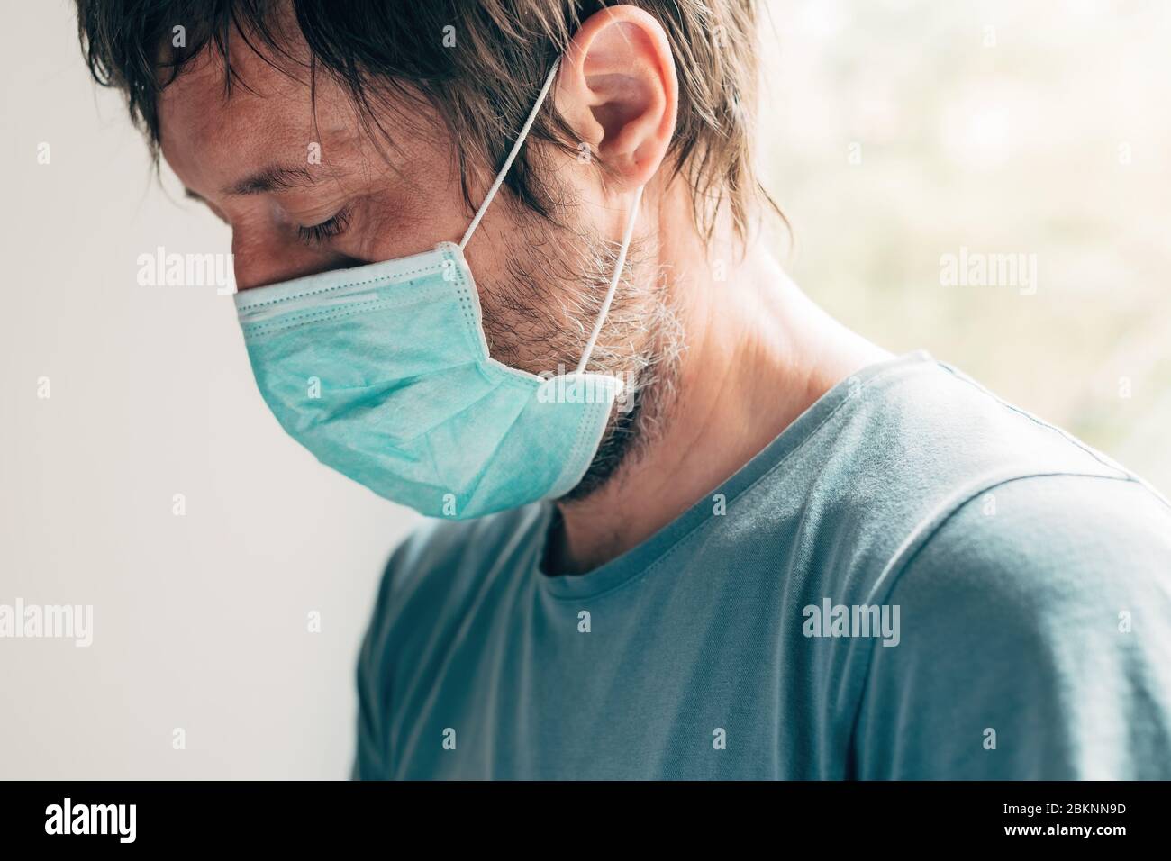 Homme déprimé avec masque de protection en quarantaine auto-isolée pendant une pandémie de virus, homme adulte debout par la fenêtre Banque D'Images