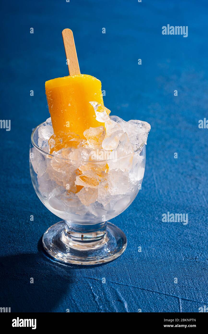 Rafraîchissement de la popsicle orange dans un verre avec de la glace sur une table bleue classique. Dessert tropical froid. Bonbons d'été. Glace orange sur un bâton. Banque D'Images
