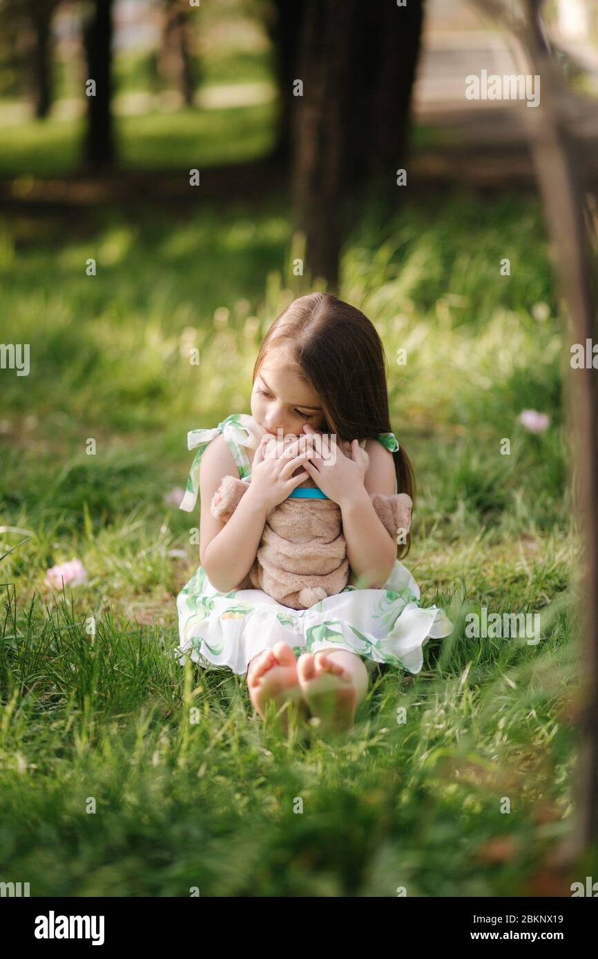 Une petite fille adorable s'assoit sur l'herbe pieds nus et l'ours en peluche. Les enfants passent du temps dans le parc, à l'extérieur. Magnifique modèle petite fille Banque D'Images