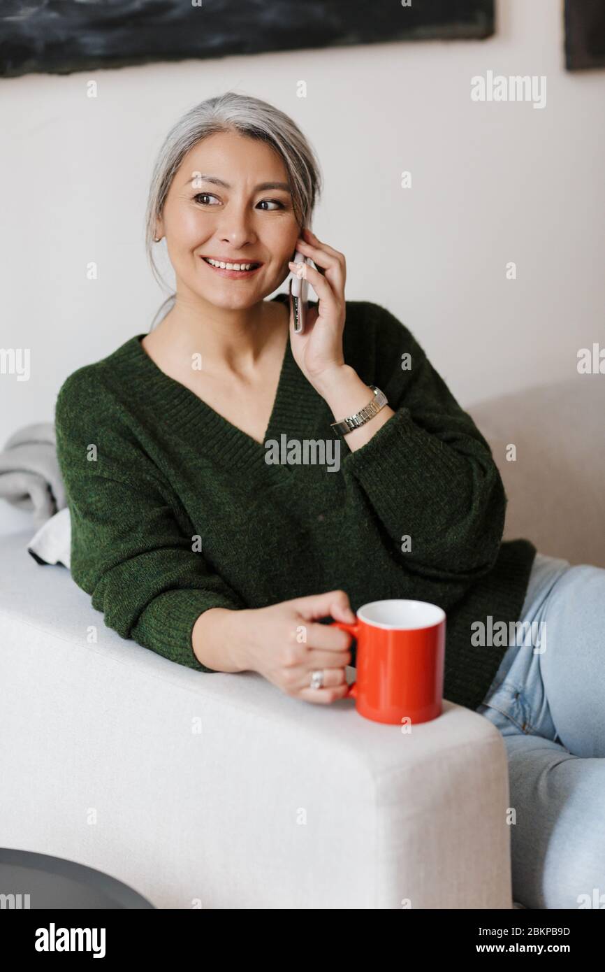 Image d'une femme asiatique adulte qui parle au téléphone portable et qui boit du café tout en étant assise sur un canapé à la maison Banque D'Images