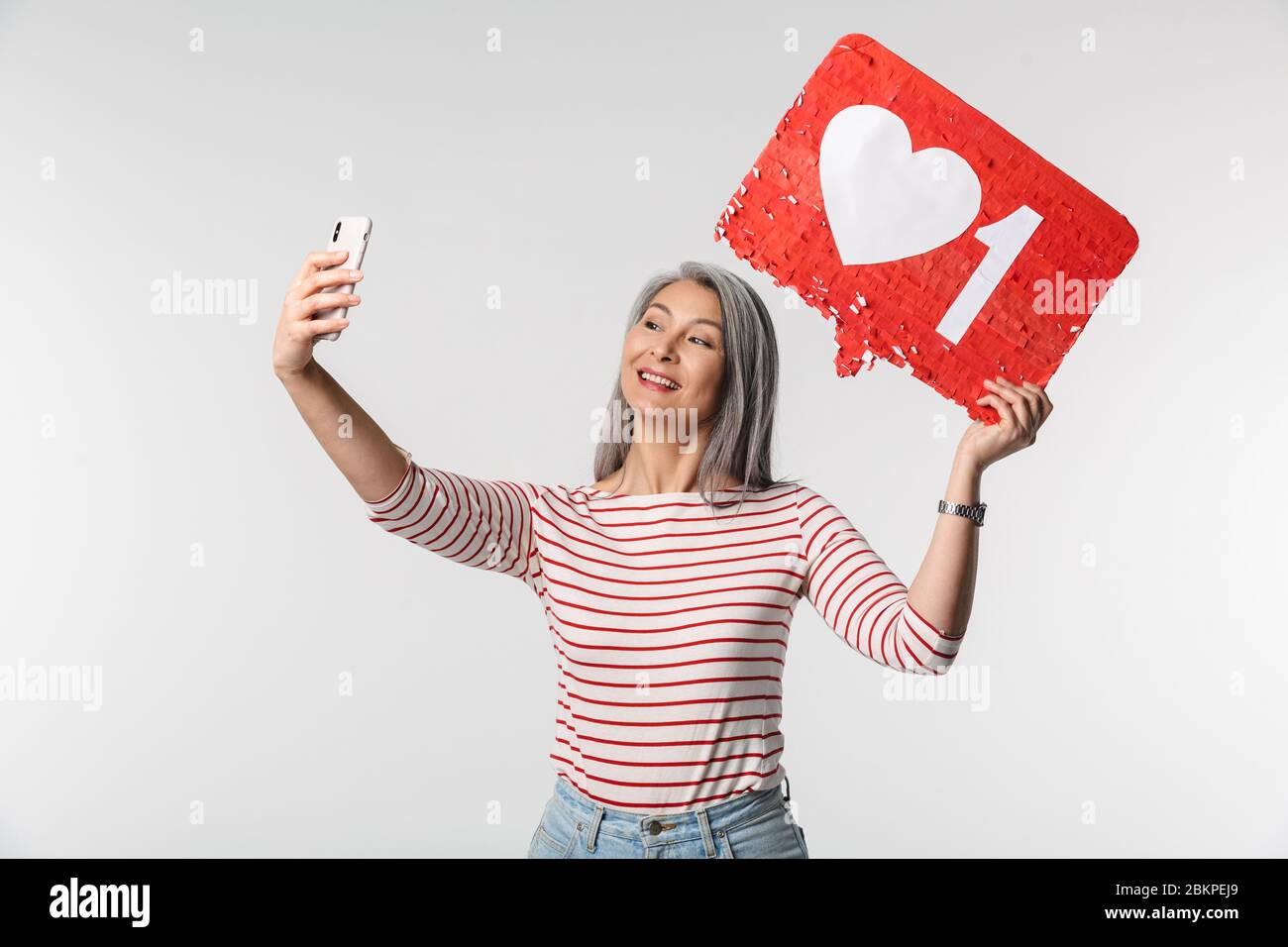 Image d'une femme adulte mature avec de longs cheveux blancs tenant un téléphone cellulaire et un symbole de coeur sur l'étiquette isolée sur fond gris Banque D'Images