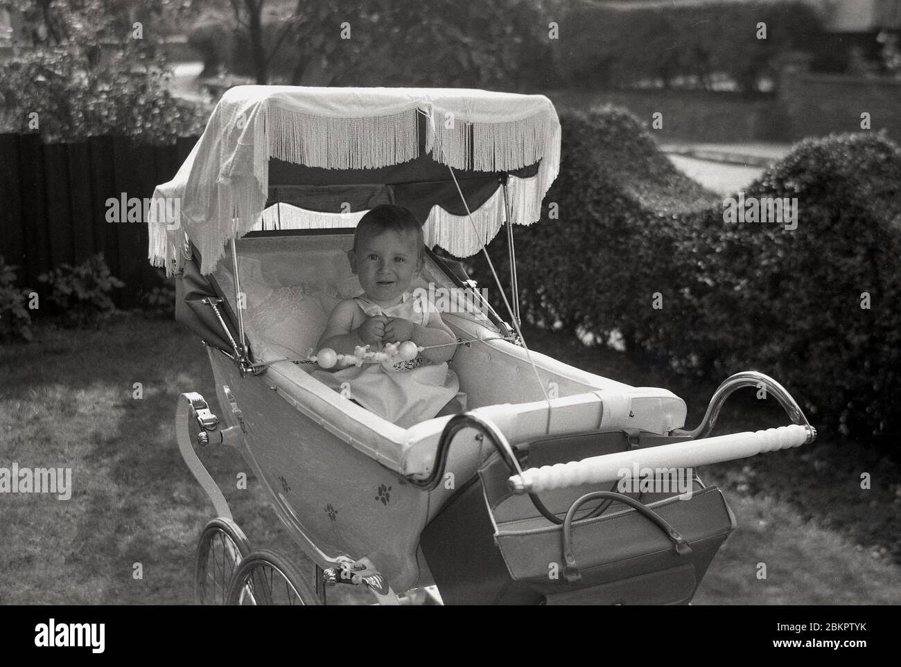 Années 1950, historique, à l'extérieur d'un jardin devant, un bébé assis heureux sous le capot ou la canopée d'une voiture traditionnelle de l'époque. Banque D'Images