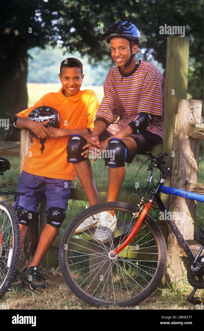 Vélos adolescents Anglais Africain Caraïbes frères américains 11 ans -15 ans portant un casque et un kit de cyclisme de protection, à l'extérieur sous un soleil d'été raillé prenant une pause pendant leurs aventures à vélo pendant les vacances scolaires d'été. Banque D'Images