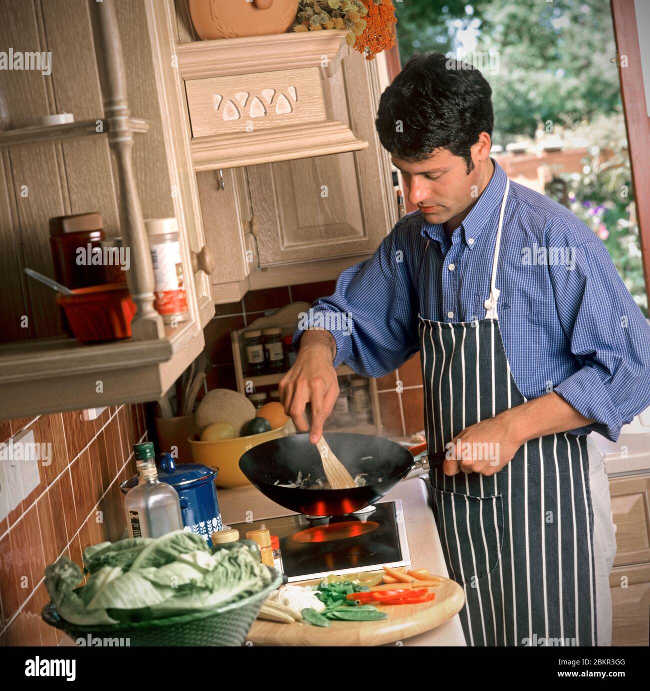 Homme des années 25-30 cuisiner un bon repas avec table de cuisson, dans sa cuisine de style des années 90, avec jardin ensoleillé en arrière-plan à travers la fenêtre ouverte de la cuisine Banque D'Images