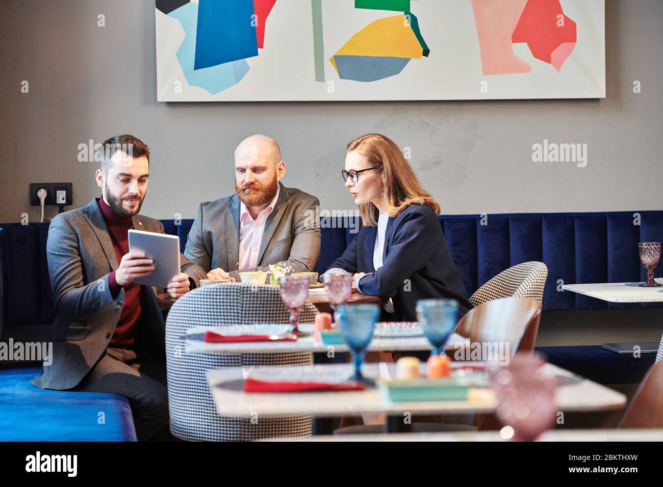 Jeune Manager barbu en veste utilisant une tablette numérique tout en présentant un projet d'entreprise à des collègues dans un café moderne Banque D'Images