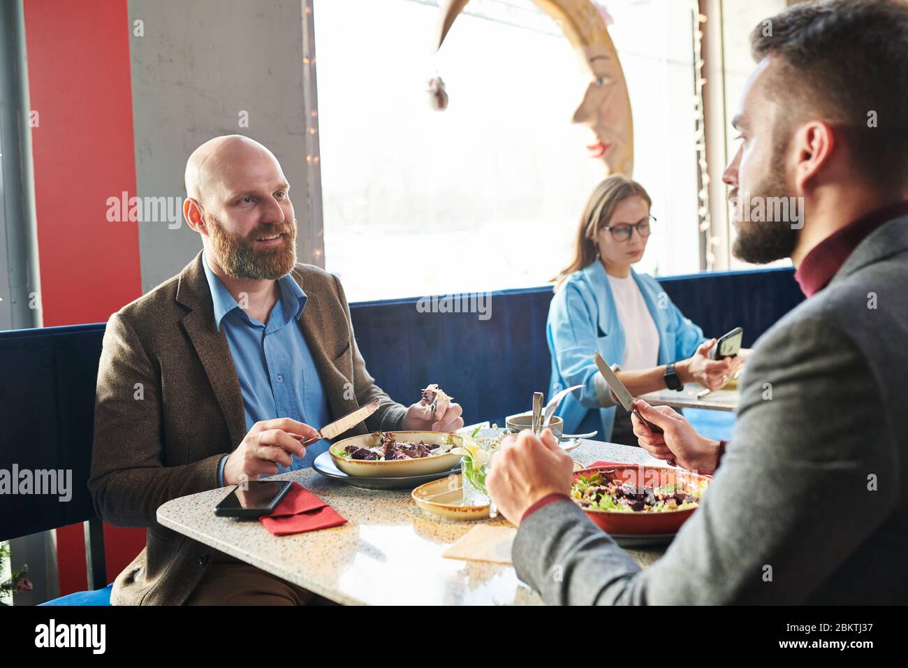 Homme d'affaires chauve souriant assis à table et en train de manger de la salade tout en discutant avec un collègue au cours d'un déjeuner au café Banque D'Images