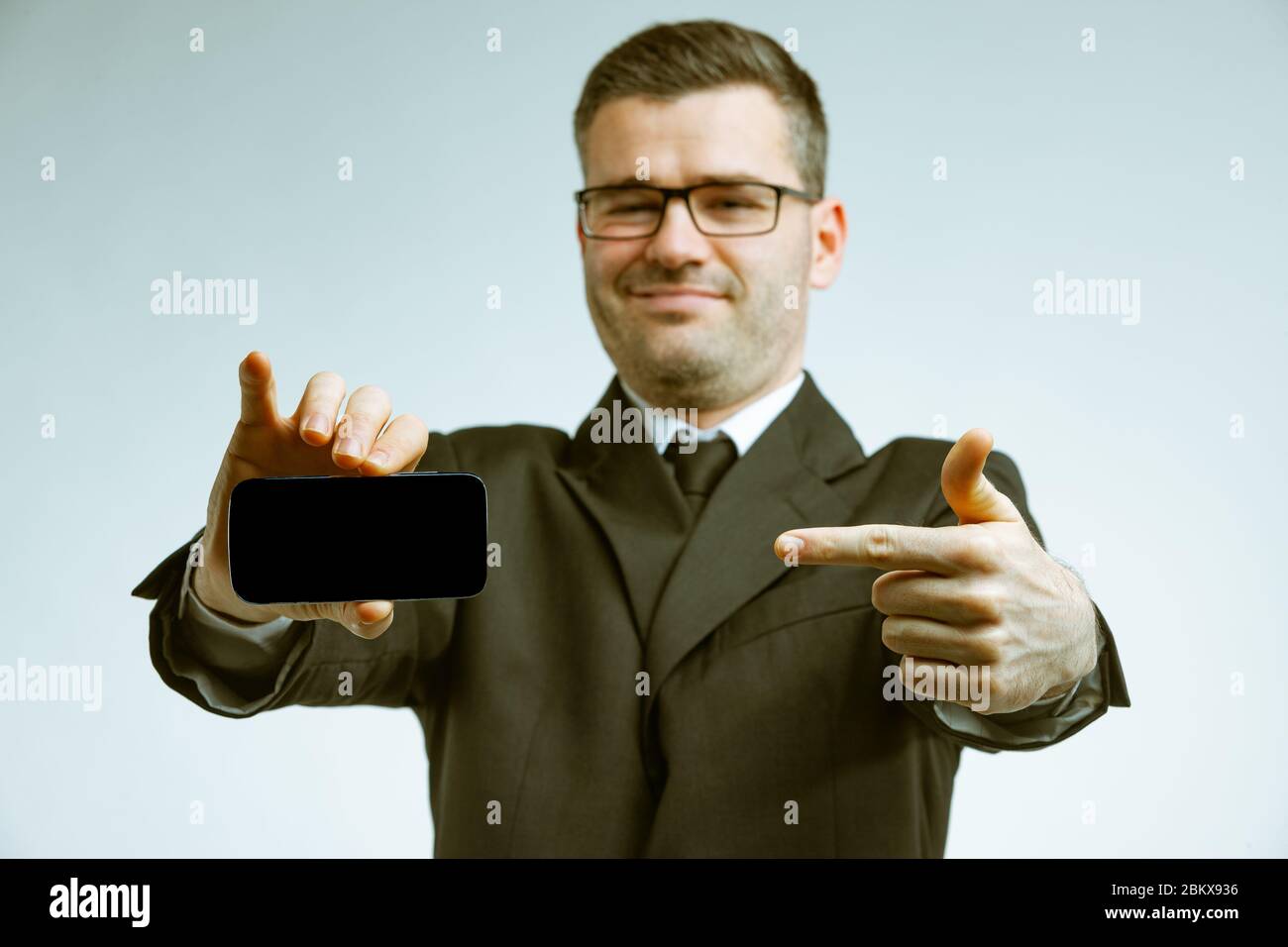 Jeune homme en costume et lunettes avec un sourire sur son visage, pointant avec son index sur l'écran noir d'un smartphone dans sa main. Fron à angle bas Banque D'Images