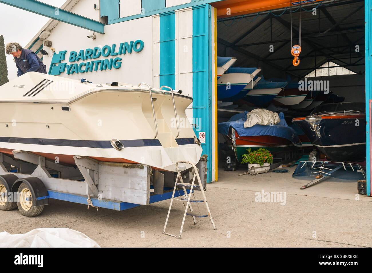 Homme effectuant des travaux d'entretien sur un bateau devant la tour de bateau Bardolino Yachting sur la rive du lac de Garde, Bardolino, Vérone, Vénétie, Italie Banque D'Images