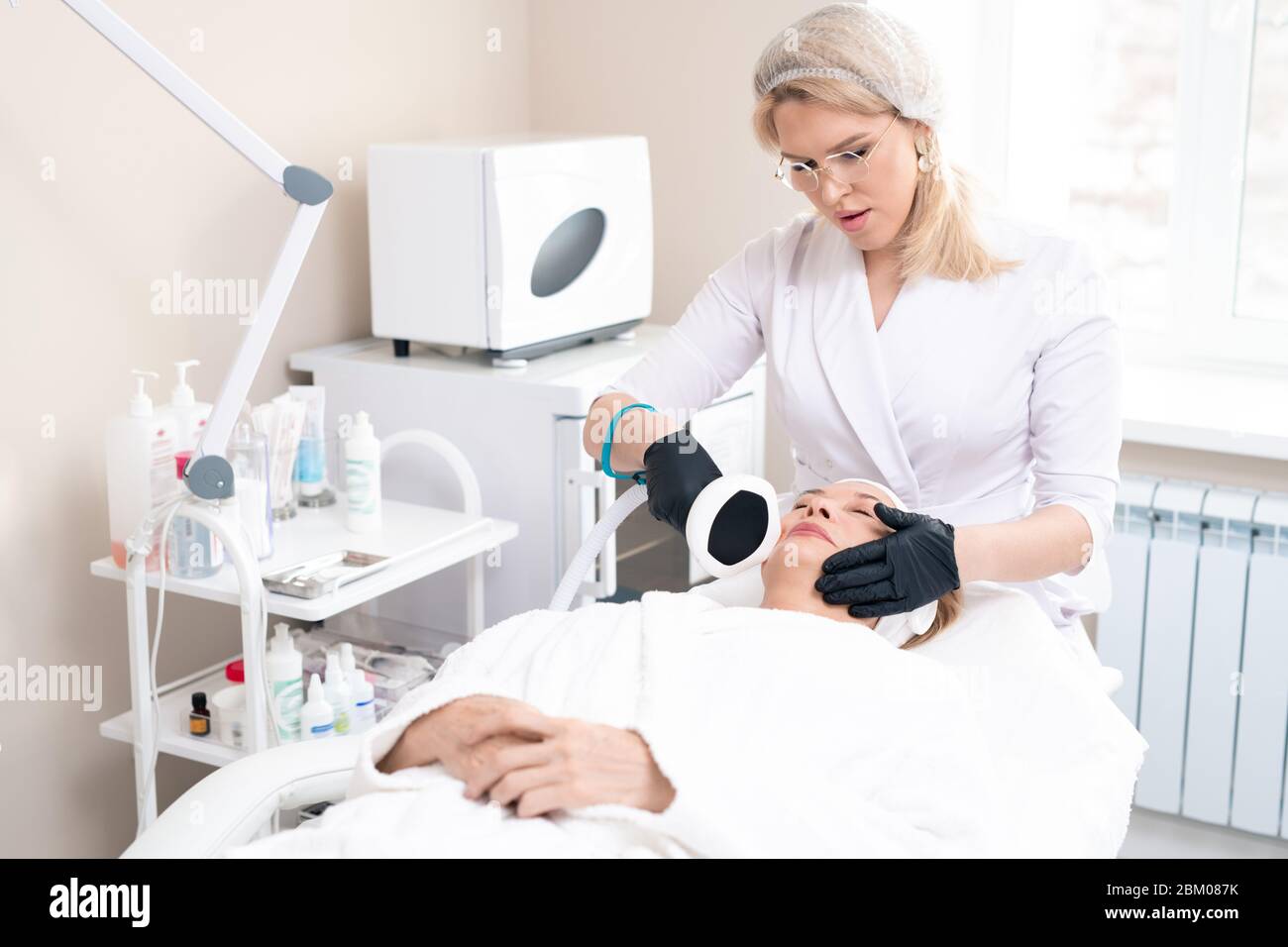 Un cosmétologue sérieux en bonnet chirurgical et gants faisant une procédure laser cosmétique à la femme mature dans le salon de beauté Banque D'Images