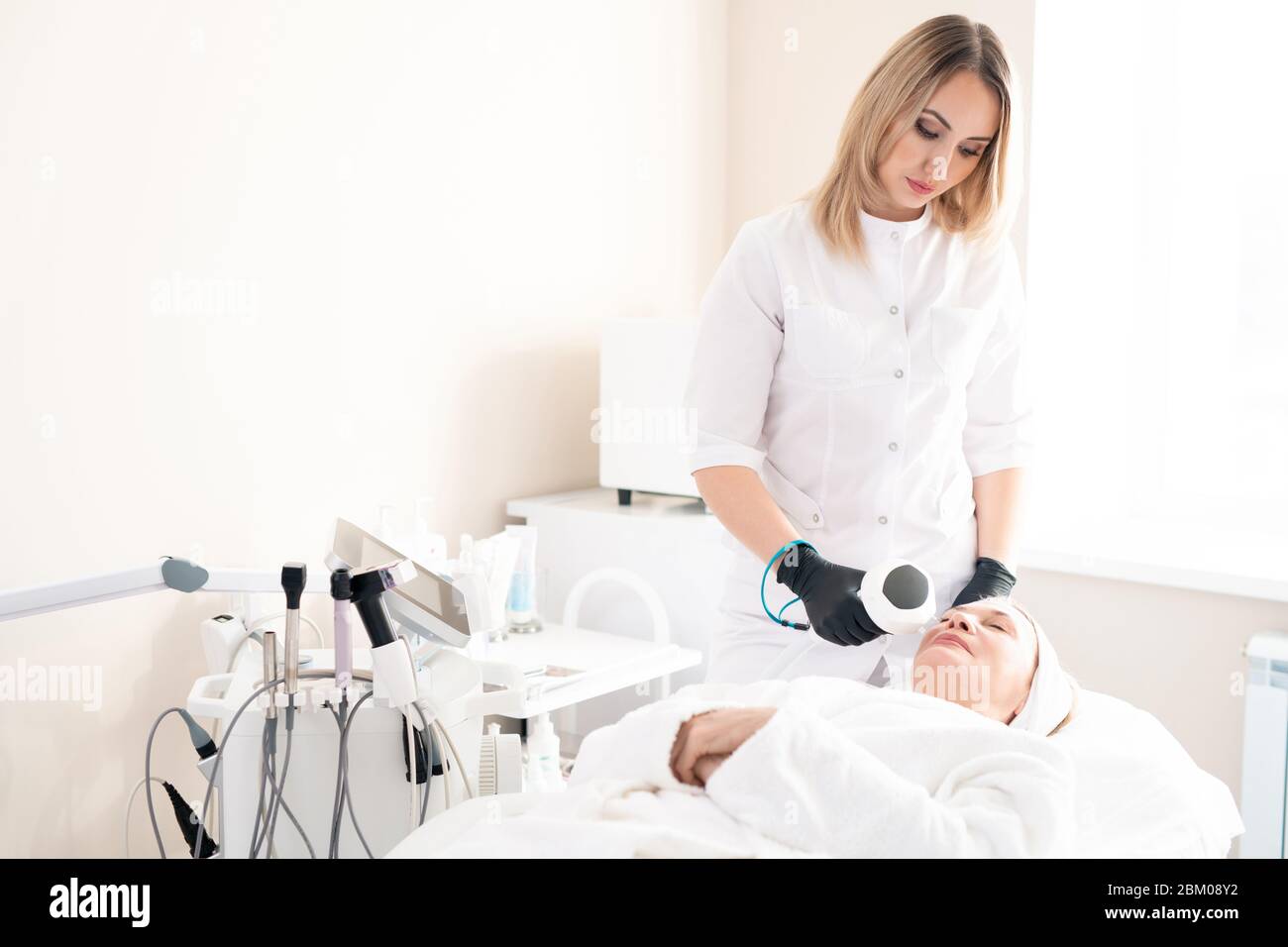 Jeune cosmétologue en gants chirurgicaux offrant un traitement laser à une femme mature sur une table d'intervention dans un salon de beauté Banque D'Images