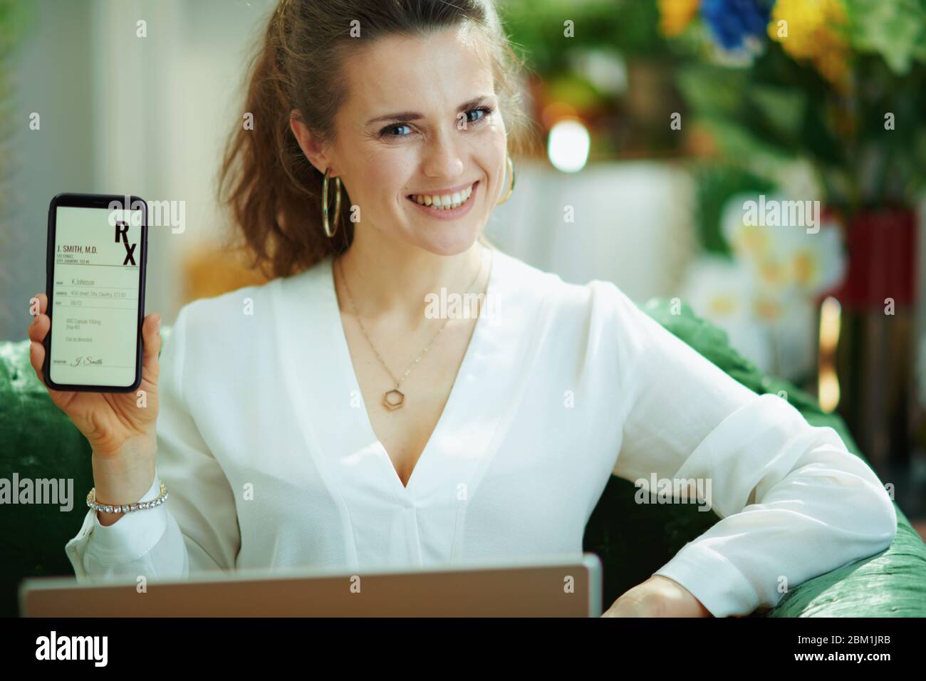 Portrait de la femme au foyer branchée heureuse dans un chemisier blanc dans la maison moderne en journée ensoleillée montrant électronique rx sur un téléphone. Banque D'Images