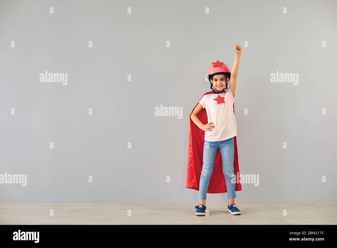 Le petit super-héros de bébé dans un casque sourit ses mains tout en se tenant sur un fond gris. Banque D'Images