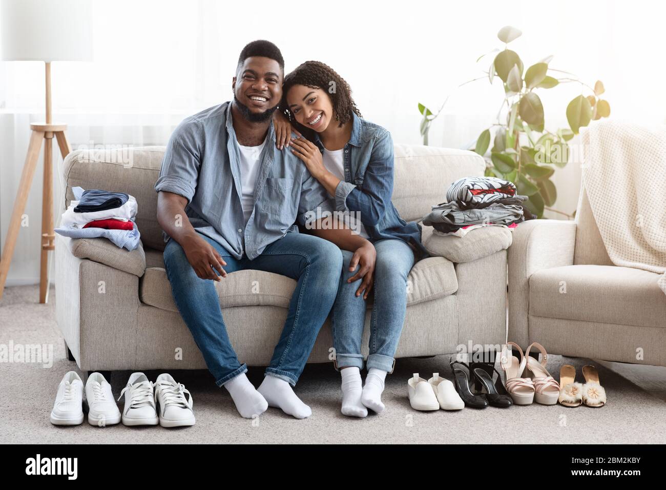 Joyeux couple noir assis sur un canapé avec des vêtements pliés et des chaussures autour Banque D'Images