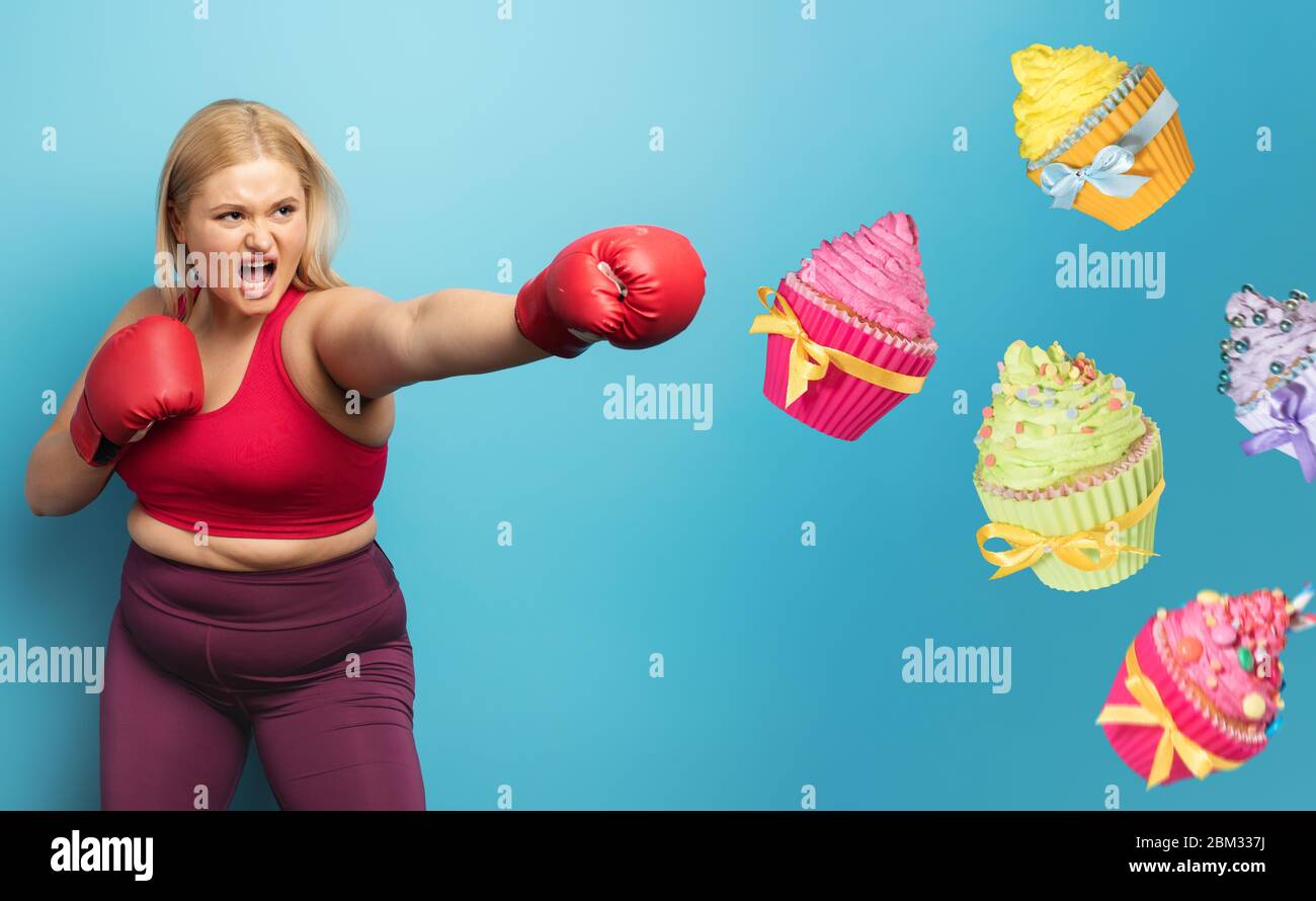 FAT girl dans la salle de fitness fait la boxe et lutte contre les cupcakes. Fond cyan Banque D'Images