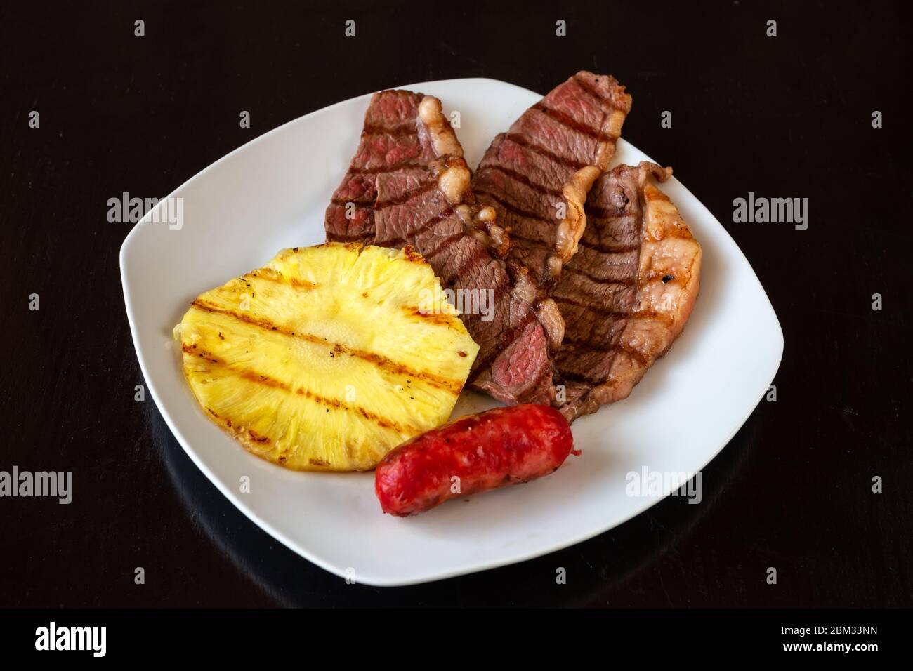Les morceaux de viande sont juteux, hachés, avec ananas et saucisse sur une assiette blanche. Arrière-plan noir foncé. Gros plan. Banque D'Images