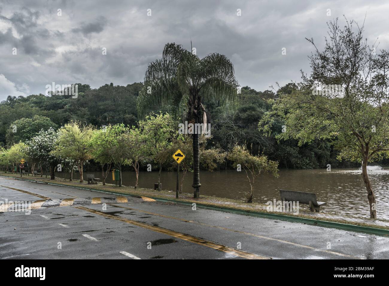 Une belle scène en journée de pluie où il est possible de voir un banc vide face à un lac, plus une forêt tropicale et un panneau de houle et un asphalte humide. Banque D'Images