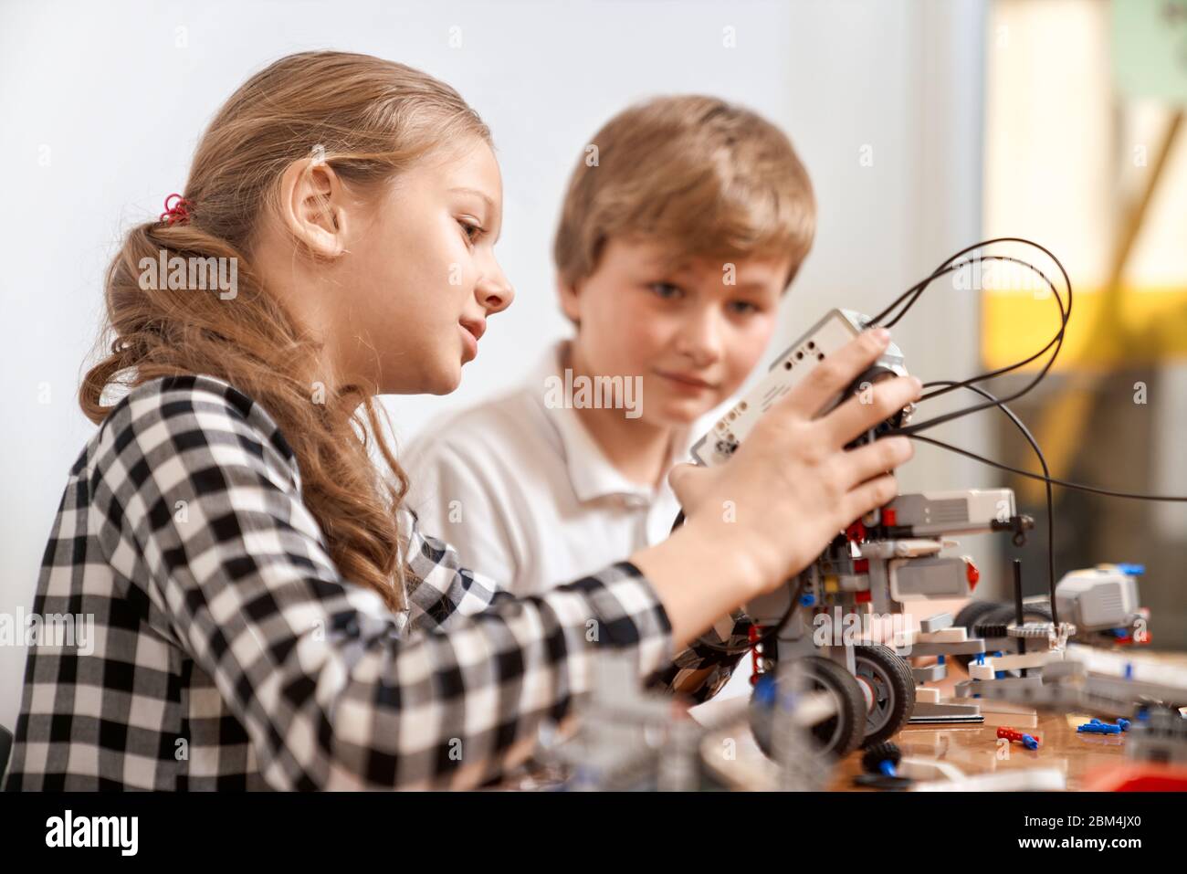 Vue latérale d'un garçon aidant une fille à créer un robot en utilisant le kit de construction pour les enfants sur la table. Des amis agréables et intéressés souriant, discutant et travaillant ensemble sur le projet. Concept de génie scientifique. Banque D'Images