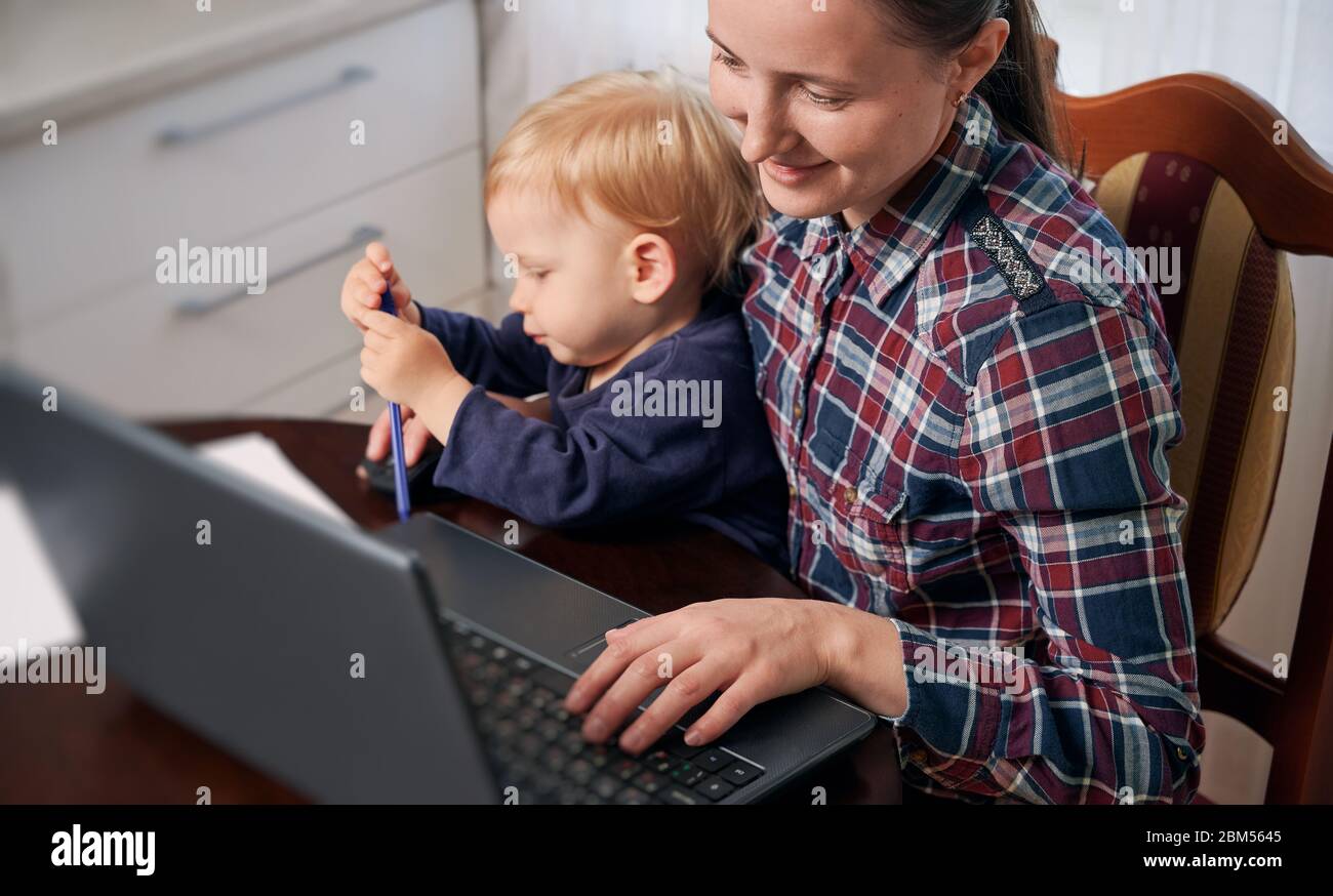 Une jeune mère reste à la maison et travaille à distance sur un ordinateur portable près de la table avec un tout-petit sur ses tours. Bureau à domicile avec des enfants en situation de pandémie mondiale. Concept Covid-19 Banque D'Images