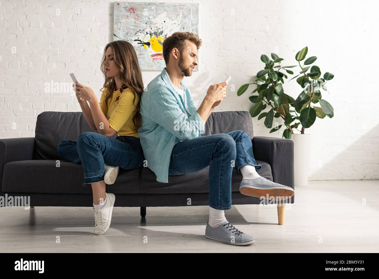 Vue latérale du couple à l'aide d'un smartphone tout en étant assis dos à dos sur le canapé Banque D'Images