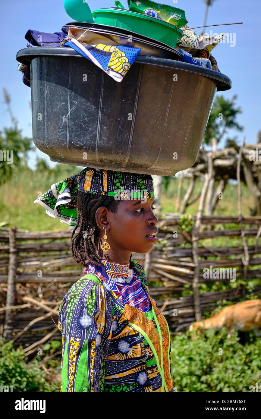 Jeune Fulani tenue de la manière traditionnelle portant sur sa tête un énorme récipient en plastique plein de pots. Banque D'Images