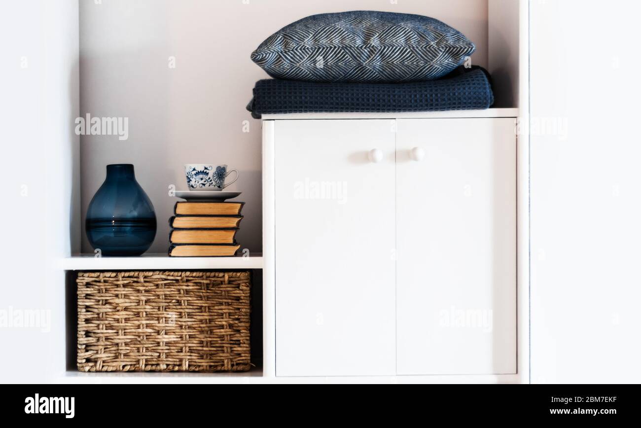 Décoration intérieure chaleureuse : oreiller, écossais, vase bleu, pile de livres, tasse de café, boîte en osier sur une étagère blanche dans la chambre. Le concept de quarantaine de Banque D'Images