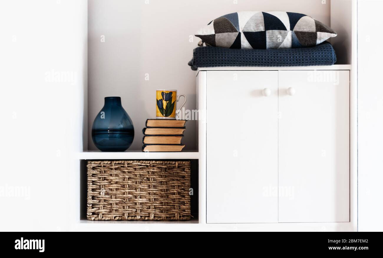 Décoration intérieure chaleureuse : oreiller, écossais, vase bleu, pile de livres, tasse de café, boîte en osier sur une étagère blanche dans la chambre. Le concept de quarantaine de Banque D'Images