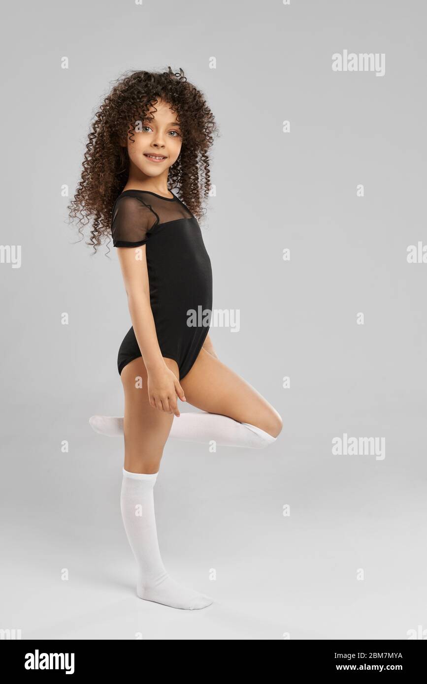 Portrait de pleine longueur d'adorable fille souriante dans des vêtements de sport noirs et chaussettes blanches de genou posant avec la jambe courbée, isolé sur fond gris studio. Petite gymnaste professionnelle féminine avec cheveux bouclés. Banque D'Images