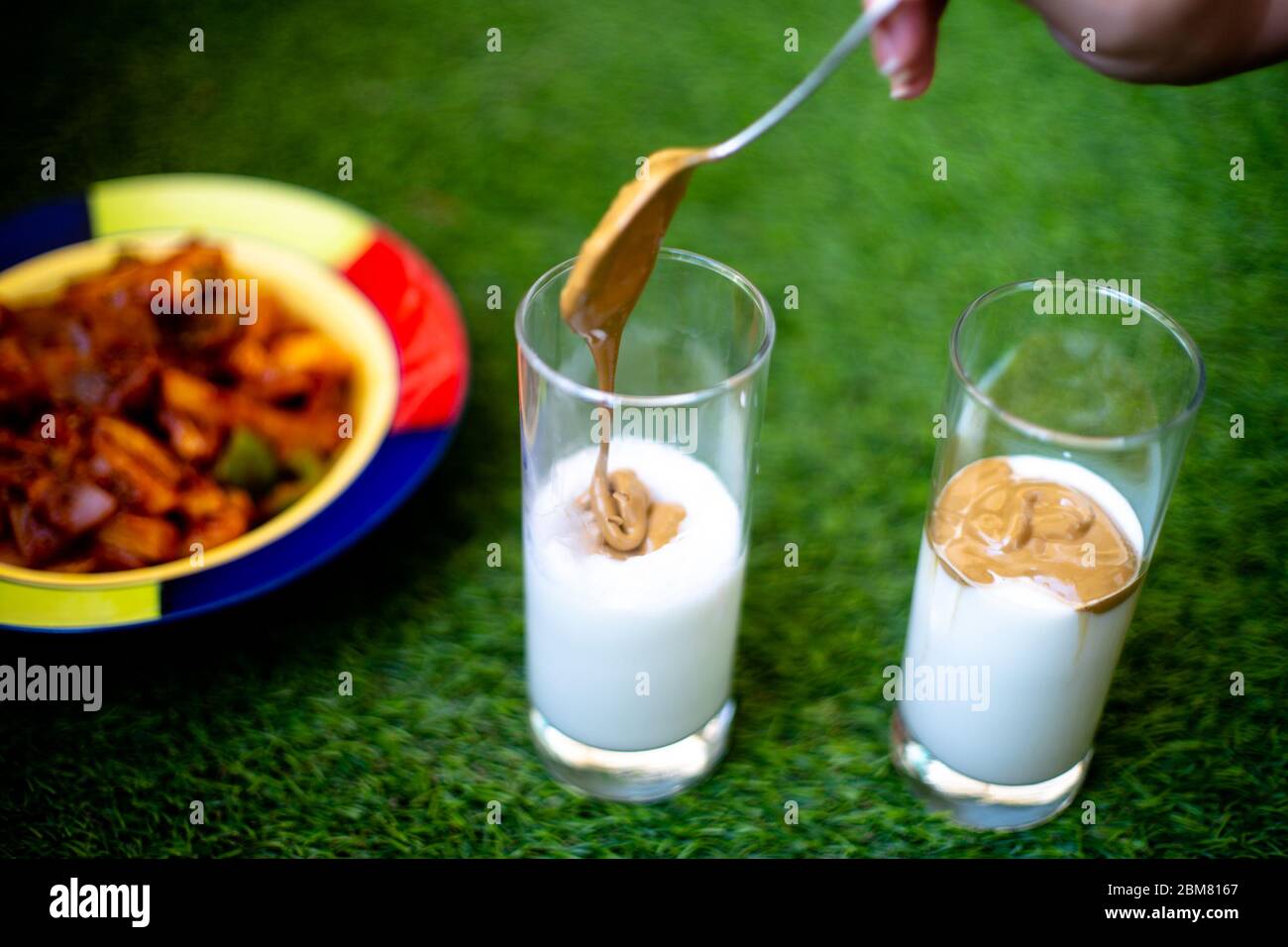 Jeune femme indienne utilisant une cuillère pour mettre du café fouetté sur du lait crème épais pour faire du café de Dalgona dans du verre transparent sur l'herbe verte Banque D'Images