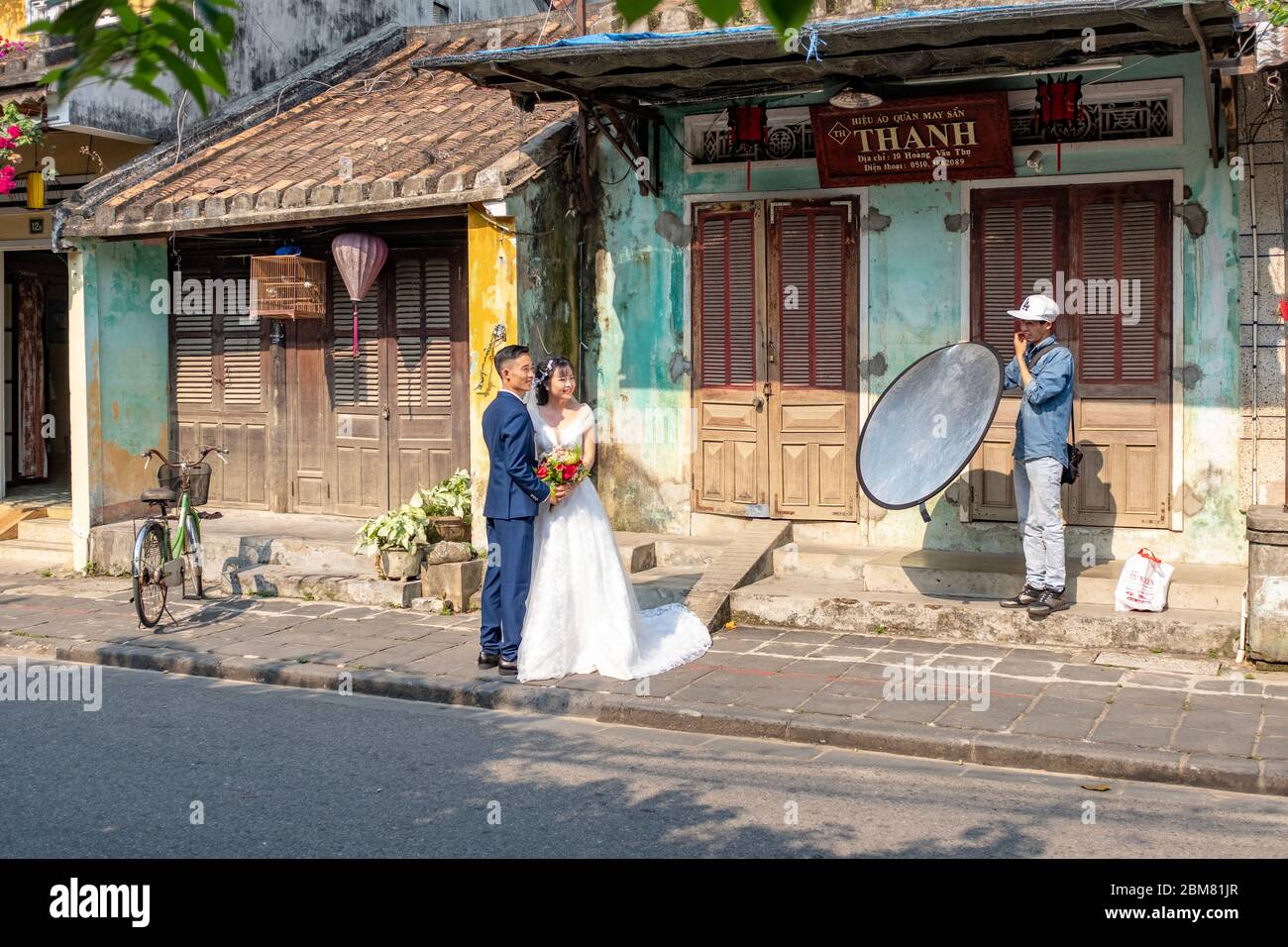 Hoi an, Vietnam - 12 avril 2018: Séance de photos et couple asiatique en tenue de mariage dans une rue de Hoi an avec un technicien tenant un réflecteur Banque D'Images