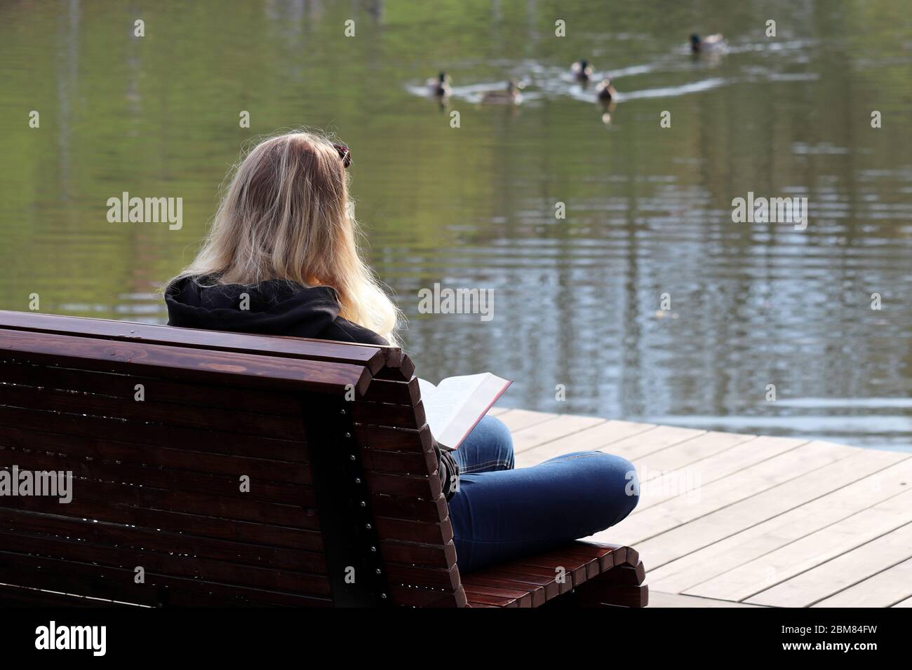 Fille blonde en Jean assis avec un livre sur banc en bois dans un parc de printemps sur fond de lac. Concept de lecture, loisirs sur la nature Banque D'Images