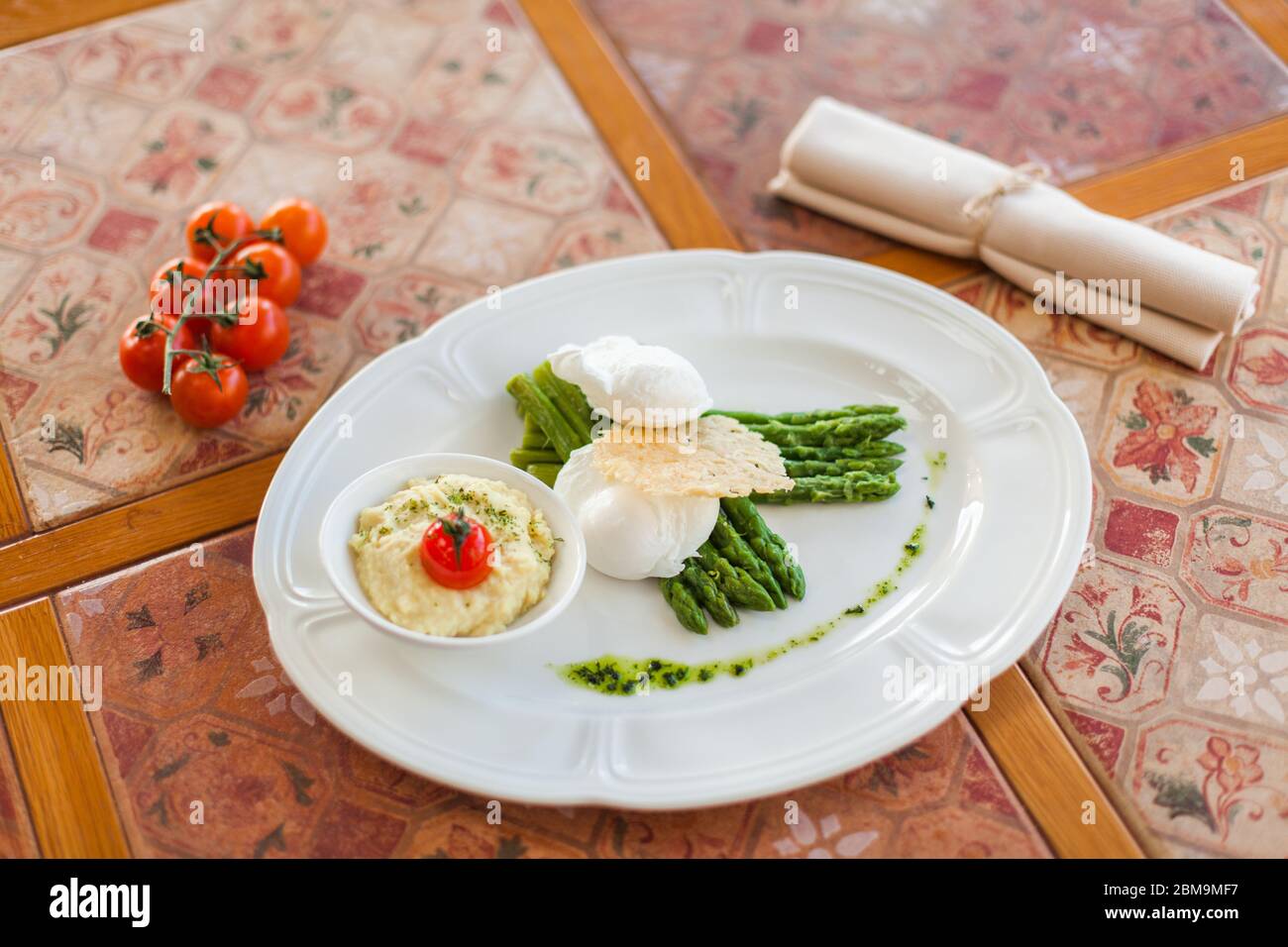 œuf poché aux asperges grillées, servi sur une assiette ovale blanche sous une croûte de fromage avec purée de pommes de terre et de tomates. table de salle à manger vintage Banque D'Images