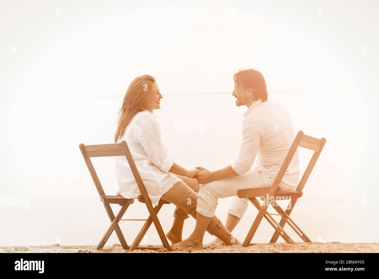 Couple mature reposant à la plage en tenant les mains. Homme et femme heureux tenant les mains et regardant l'un l'autre tout en étant assis sur une plage de sable sur fond de mer Banque D'Images