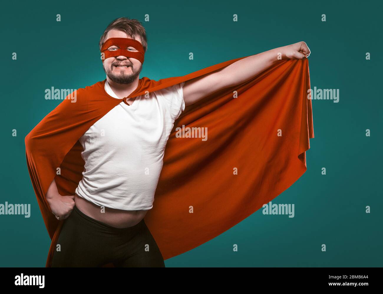 Drôle Super Hero Man prêt à voler Save World. Sourire Homme en masque et Superson Rouge Costume tenant son manteau dans sa main posant sur le vert de Gascogne Banque D'Images