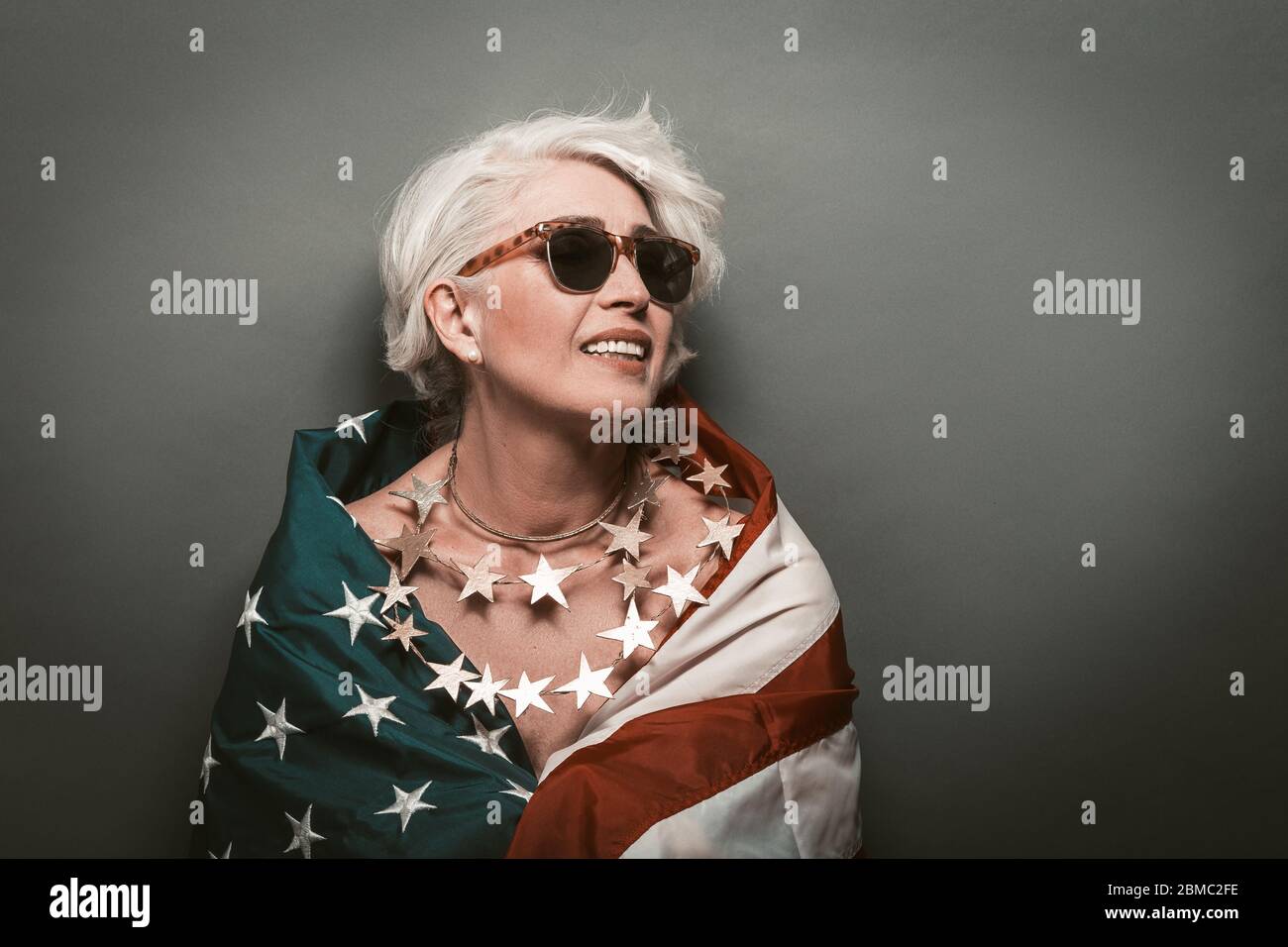 Bonne femme mature portant le drapeau des états-unis. Belle dame âgée en perles d'étoiles et lunettes de soleil sourires tortueuse regardant l'espace de copie à droite contre Banque D'Images