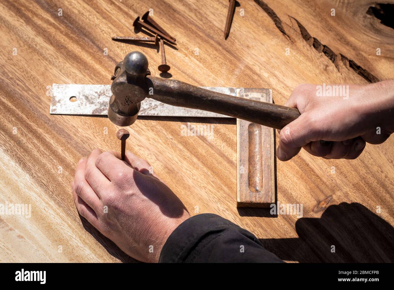 Main d'un menuisier tenant un marteau à panne de balle pour entraîner un vieil ongle rouillé dans un morceau de bois. Outil de mesure en arrière-plan. Bricolage, authentique maki Banque D'Images