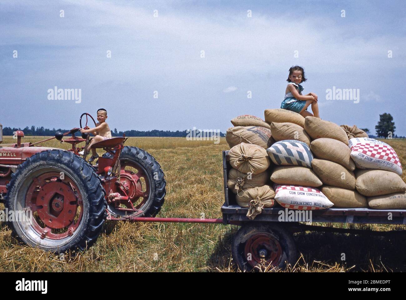 Deux enfants jouant au soleil sur un tracteur et une remorque dans la ferme familiale, aux États-Unis, au début des années 1950. Les sacs rouges et bleus de la remorque contiennent le concentré de Chow Chow, un supplément pour l'alimentation animale. Banque D'Images