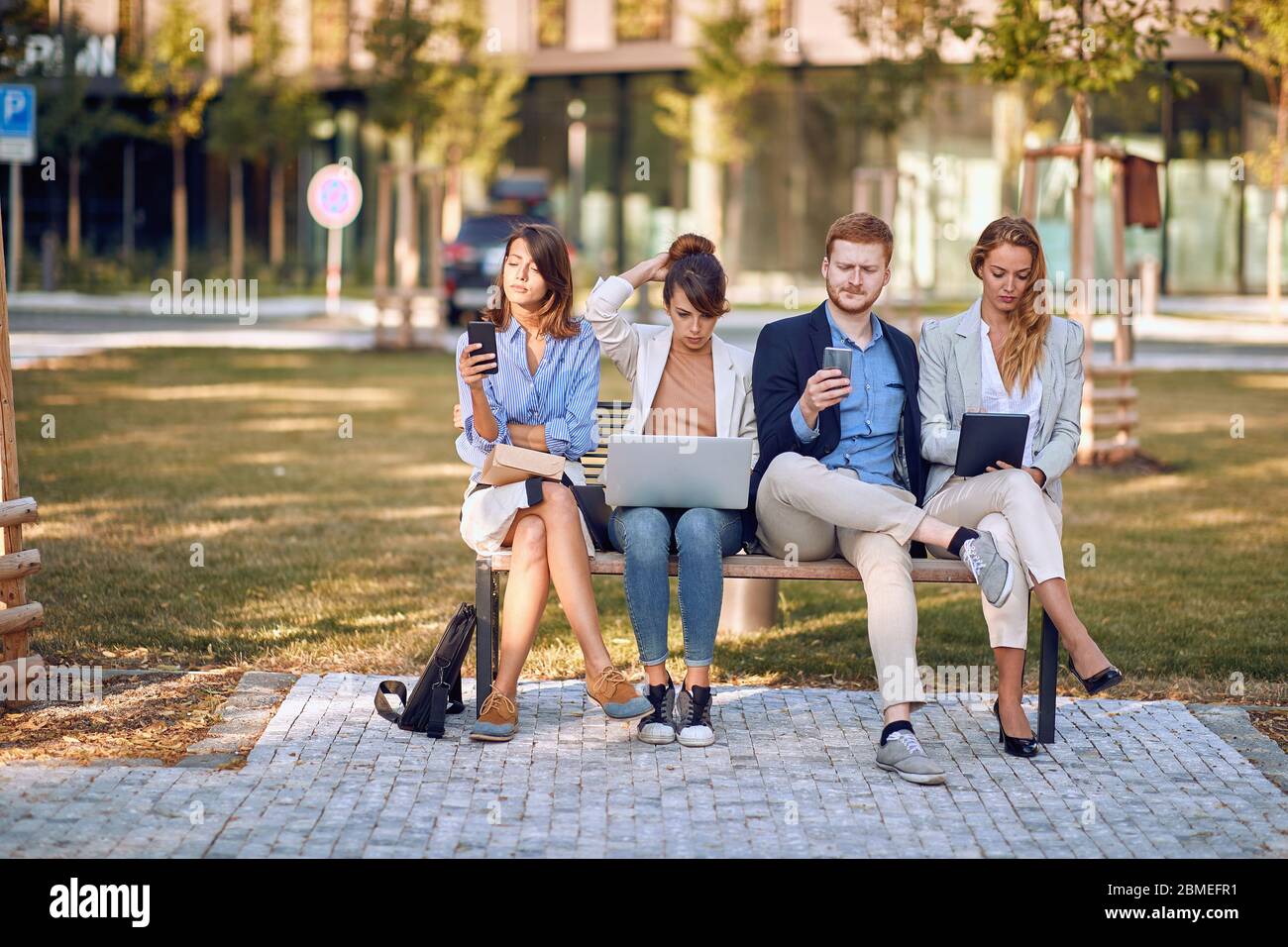 Les gens d'affaires sur le banc au parc, travaillant sur un ordinateur portable, message texte. Jeunes employés à l'extérieur ensemble. Banque D'Images