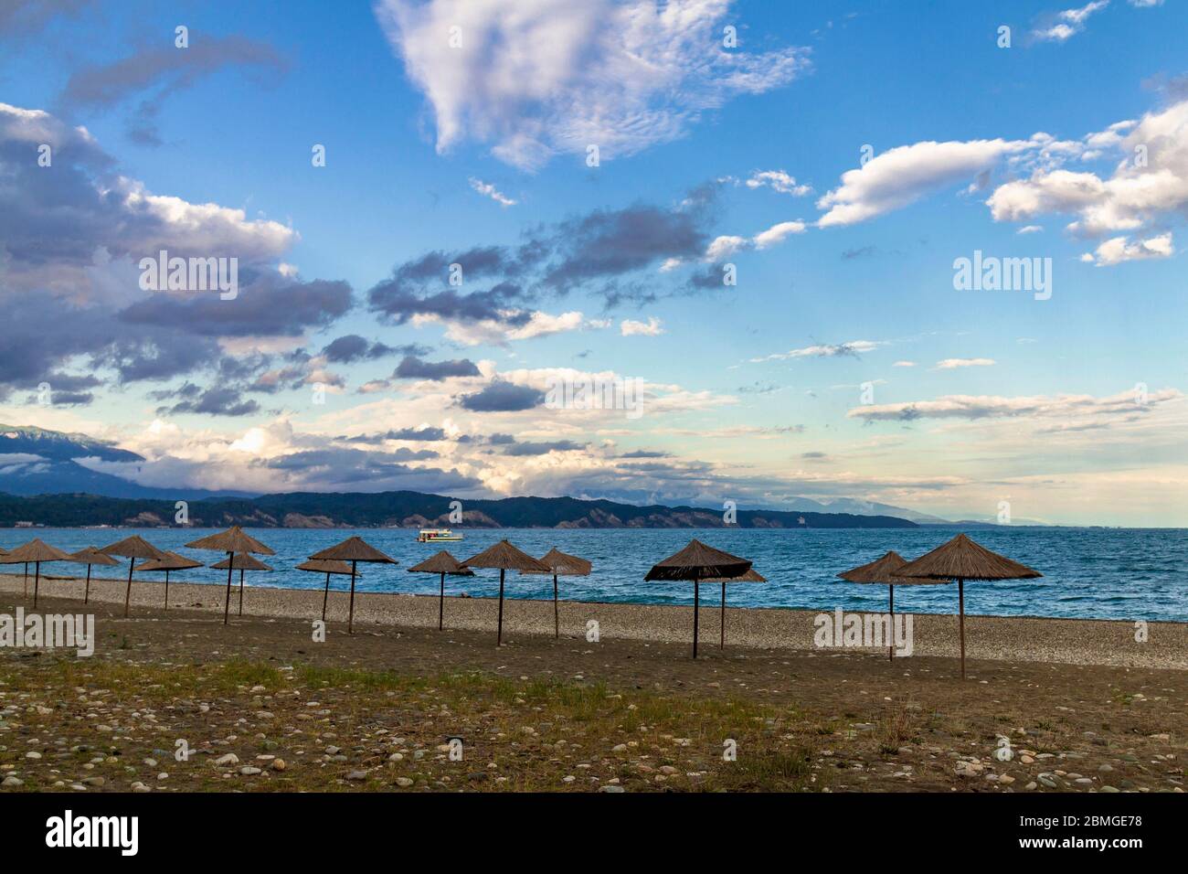 Parasols de plage de paille sur la côte vide de la mer Noire en Abkhazie, Pitsunda au coucher du soleil. Plage de galets, montagnes, mer, ciel avec nuages Banque D'Images