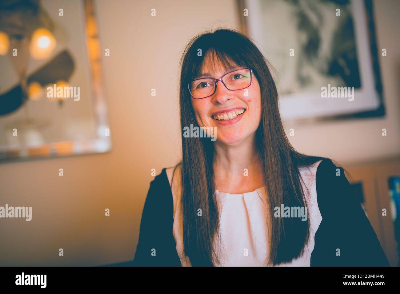 Vue de face de la femme britannique ordinaire en lunettes souriant, femme isolée à l'intérieur, salle de séjour du Royaume-Uni. Banque D'Images